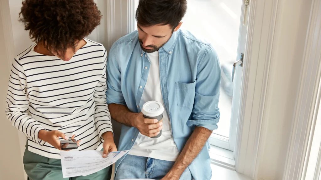 Top view of multiethnic managers discuss marketing report, work together, check information on mobile phone drink takeaway coffee stand against window wall. Unshaven man and his assistant.
